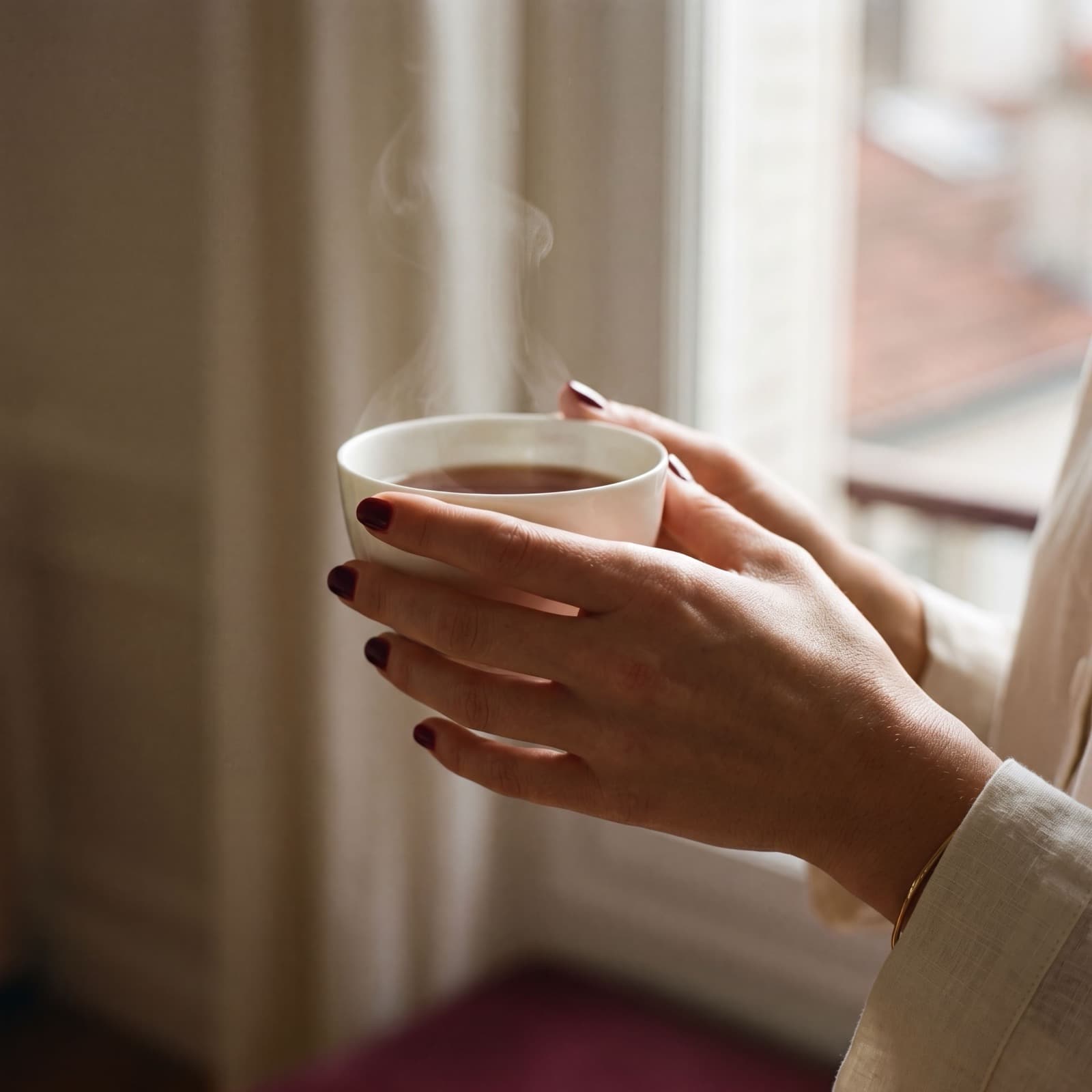 Mains aux ongles bordeaux tenant une tasse de thé fumante près d'une fenêtre